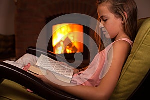Young girl in confortable armchair reading in front of fireplace photo