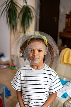 A young girl with curly hair and a lime headband smiles gently in a home setting, exuding warmth and innocence.