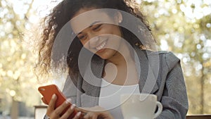 Young girl with dark curly hair using phone while sitting in cafe, outdoors.