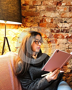 A young girl with dark long hair and glasses sits in a cafe and reads notes in a notebook. A young woman in a gray sweater sits on
