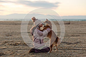 A young girl with a dog in nature. Kid girl hugging a shiba inu dog on the beach at sunset in Greece in winter