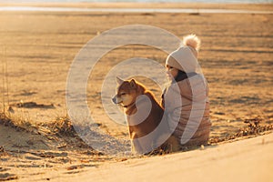 A young girl with a dog in nature. Kid girl sitting with a shiba inu dog on the beach at sunset in Greece in winter