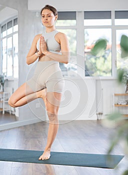 Young girl doing hatha yoga in modern studio, performing balancing asana Vrikshasana with hands clasped in namaste