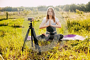 A young girl is doing yoga, sitting in the lotus position in nature and recording a video lesson about yoga. Yoga online