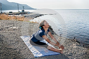 Young girl is doing yoga sitting on a stony sea coast in summer morning.