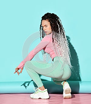 Young girl with dreadlocks hairstyle in sexy sportswear and sneakers sitting backwards and practicing booty dance