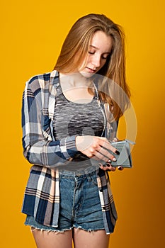 Young girl dressed in denim shorts and a shirt, holding a gray purse, pulls out money on a yellow background