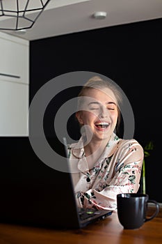 Young girl dressed in pajamas is looking at laptop screen and laughing while working or studying on a laptop at home in