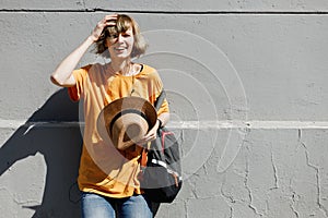 Young girl with dressed in a yellow t-shirt holds a straw hat in her hand stands leaning on a gray building in a city