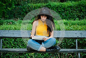 Young girl dressed in yellow and wearing denim overalls sitting on a bench while writing or drawing in a notebook, park of autumn