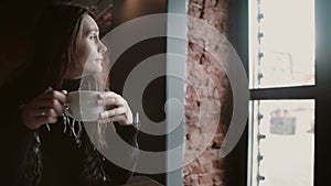 Young girl drinking tea from a mug and looking out the window sitting in modern loft dining 4k