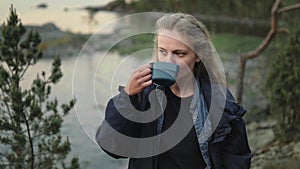 A young girl is drinking tea in nature. A tourist is drinking coffee near the lake.