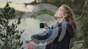 A young girl is drinking tea in nature. A tourist is drinking coffee near the lake.