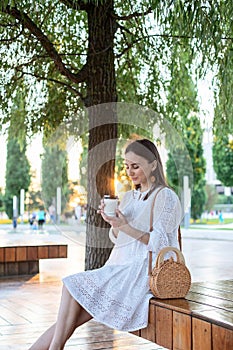 A young girl drinks coffee in the park sitting on a bench. In the rays of the setting sun, he relaxes and rests