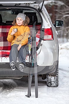 Young girl drinks hot tea after skiing while sitting in trunk of suv car. Winter activity