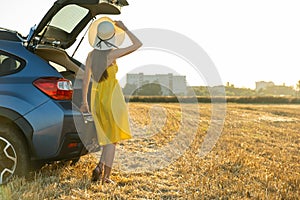 Young girl driver in yellow summer dress and straw hat standing near a car enjoying warm summer day at sunset. Travel and vacation