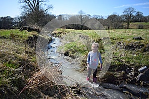 A young girl enjoying herself wading in a stream, wearing Wellington boots and the weather is sunny.