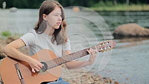 Young girl of European appearance playing an acoustic guitar on the shore of the lake, hobby concept, musical creativity