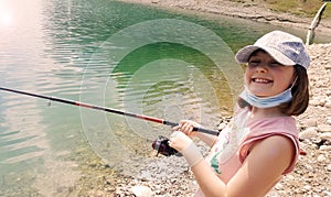 A young girl fishing smiling in a lake during conavirus time with surgical mask hanging on the neck