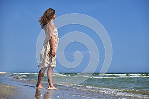 Young girl in a flower sundress stands on the seashore and looks into the distance