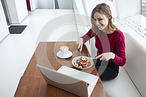 Young girl freelancer sits in a cafe with a laptop, drinks coffee and has breakfast, a student eats in the morning with a computer