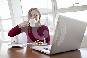 Young girl freelancer sits in a cafe with a laptop, drinks coffee and has breakfast, a student eats in the morning with a computer