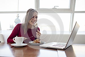 Young girl freelancer sits in a cafe with a laptop, drinks coffee and has breakfast, a student eats in the morning with a computer