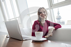 Young girl freelancer sitting in a cafe with a laptop and drinking coffee, woman use a computer near the window