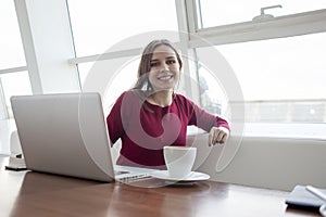 Young girl freelancer sitting in a cafe with a laptop and drinking coffee, woman use a computer near the window