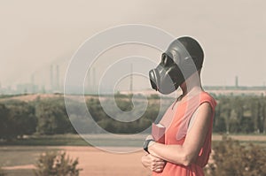 Young girl in a gas mask in a red dress with a book in her hands against the background of smoking factory chimneys on the horizon