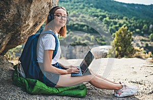 Young girl with glasses and backpack enjoying vacation in mount using headphones and laptop in nature, traveler woman planning