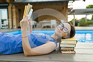 Young girl in glasses near the pool with a pile of books, reading book. Education, summer, knowledge.