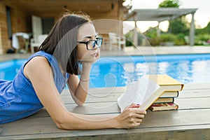 Young girl in glasses near the pool with a pile of books, reading book. Education, summer, knowledge.