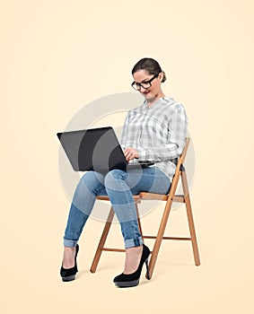 A young girl in glasses, shirt and jeans is sitting on a chair and working on a laptop, on a bright yellow background