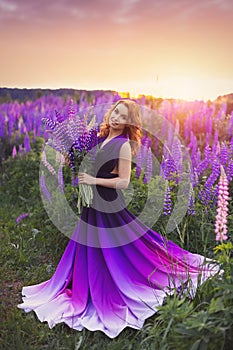 A magical portrait of a young girl in a gradient dress in a purple-pink lupine field at sunset.