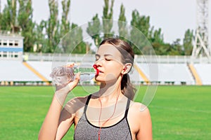 Young girl in gray sportswear drink water and listening music after teaining. woman standing and drinking with bottle at stadium.