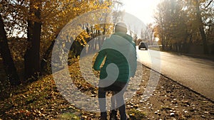 Young girl in green jacket rides bicycle in sun, on side of an asphalt road, in an autumn park, against a background of