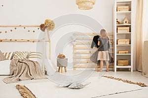 Young girl hanging clothes on wooden hanger in white Nordic style bedroom interior with home-shape bed, two pillows placed on car