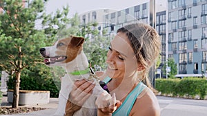 A young girl with a happy smile stands outdoors in a park area of the city, holds pet, a Jack Russell Terrier dog, in