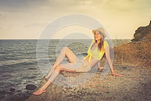 Young girl in the hat, shorts and a short shirt is sitting on a wild stony sea coast.