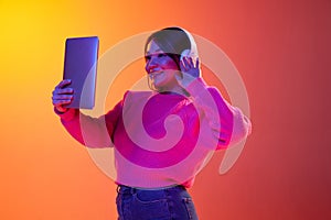 Young girl in headphones having online video call on tablet over gradient orange background in neon light. Concept of