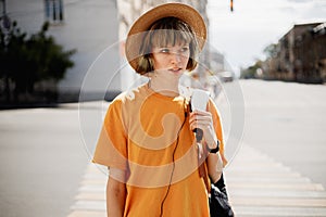 Young girl with headphones in a yellow T-shirt and a straw hat walks with a backpack along a city street on a summer