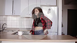 Young girl in her 20`s dancing in the kitchen with headphones in her ears, takes a cup from the table.