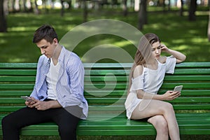 Young girl and her boyfriend with smartphones ignoring each other during dull date at park