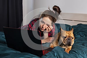 Young girl and her dog french bulldog working in bed at a laptop during quarantine