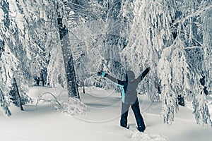 Young girl hiker in ski suit looks at tall snowy fir trees covered with snow in the forest mountains landscape. Winter holiday and