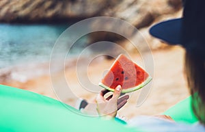 Young girl hipster relax on beach coast and holds in her hand a slice of red fresh fruit watermelon on blue sea background, woman