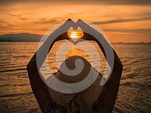 Young girl holding hands in heart shape framing setting sun at sunset