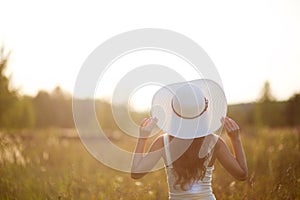 Young girl holds a big bright hat by the field and looks at the sunset. Backlight, selective focus. Young woman in hat