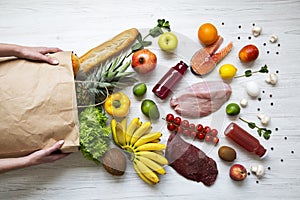 Young girl holds full paper bag of groceries on white wooden background. From above. Top view.